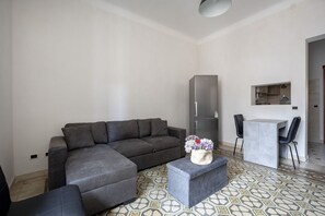 Ein modernes Wohnzimmer mit einem grauen Ecksofa, einem kleinen Tisch mit zwei Stühlen, einem grauen Kühlschrank und einer durch eine offene Tür sichtbaren Küchenzeile.