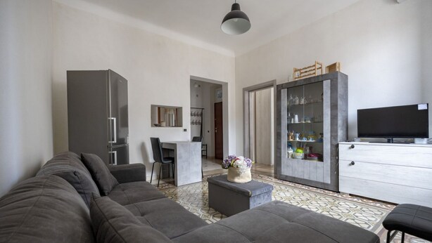 En moderne stue med en grå sofa, et lille bord med en vase, et køkkenområde med et fjernsyn og et spiseområde med stole.