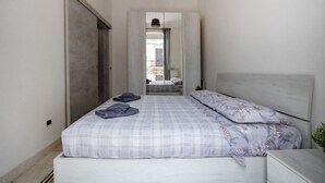 Et soveværelse med seng, natbord, garderobe og udsigt til et andet rum gennem en åben dør.