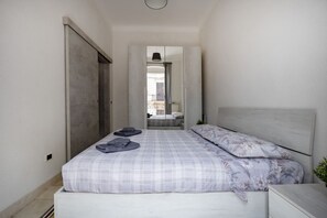 Une chambre à coucher comprenant un lit, une table de chevet, une armoire et donnant sur une autre pièce par une porte ouverte.