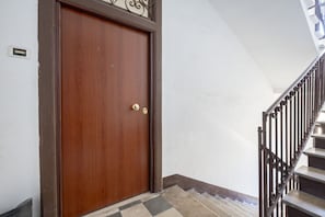 Eine Holztür mit Messinggriff, ein Metalltreppenhaus mit Geländer und ein Fliesenboden.