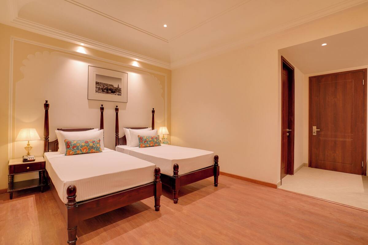 Quarto de hotel com duas camas, uma mesinha de cabeceira com abajur, um quadro na parede e uma porta de madeira.
