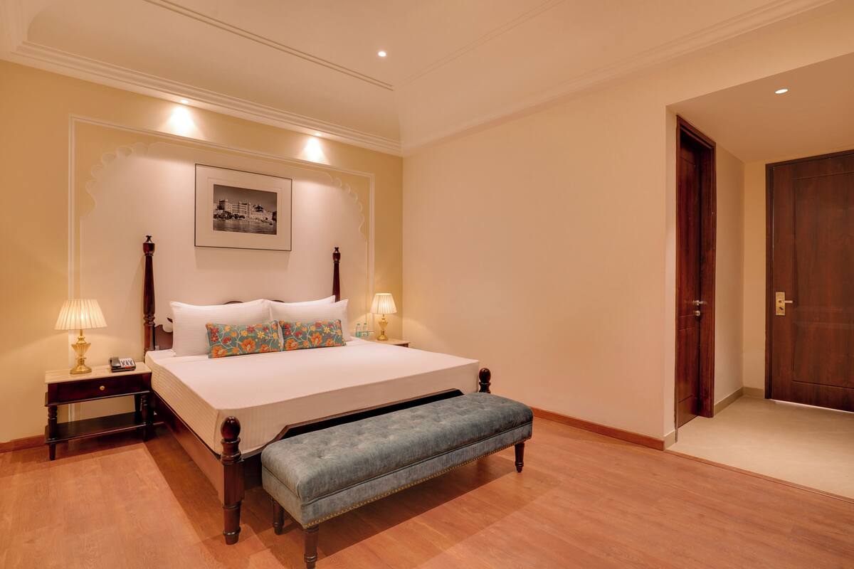 Quarto com cama grande, abajures, um banco, um quadro na parede e uma porta de madeira.