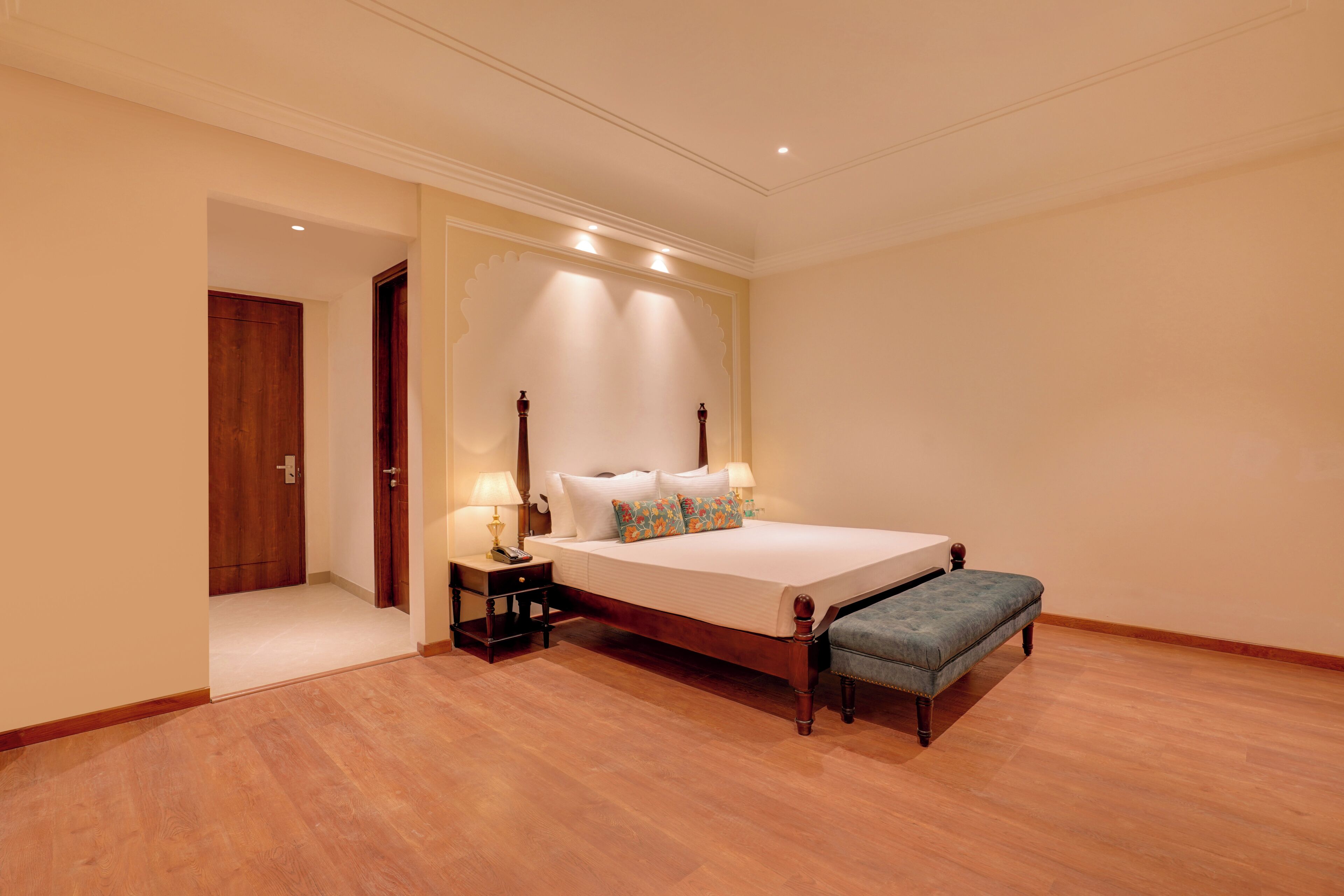 Un dormitorio con piso de madera, una cama con ropa blanca, una mesita de noche con lámpara y una puerta a otra habitación.