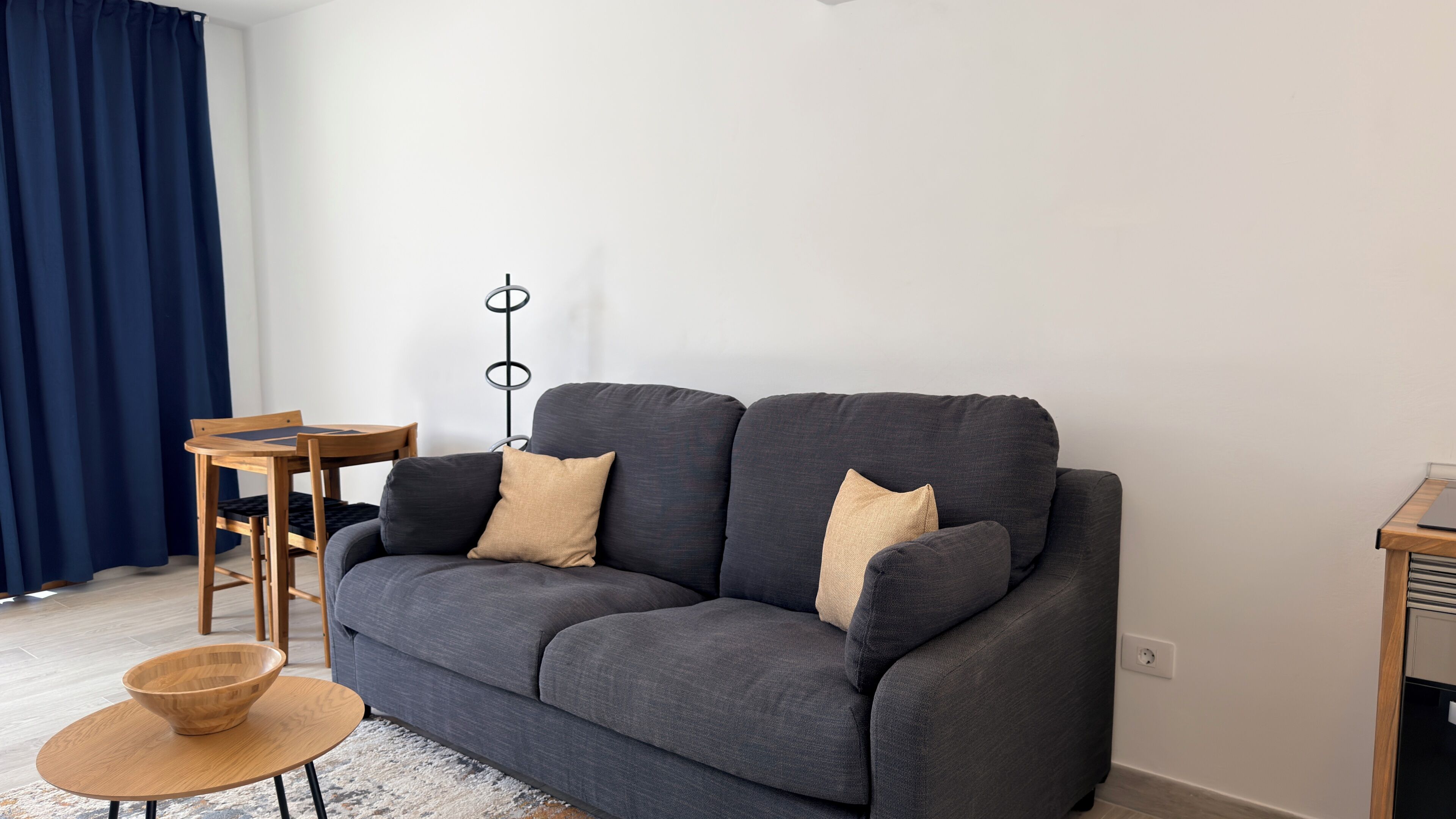 Een moderne woonkamer met een grijze bank, een klein houten bijzettafeltje, een ronde houten salontafel en een vloerlamp.