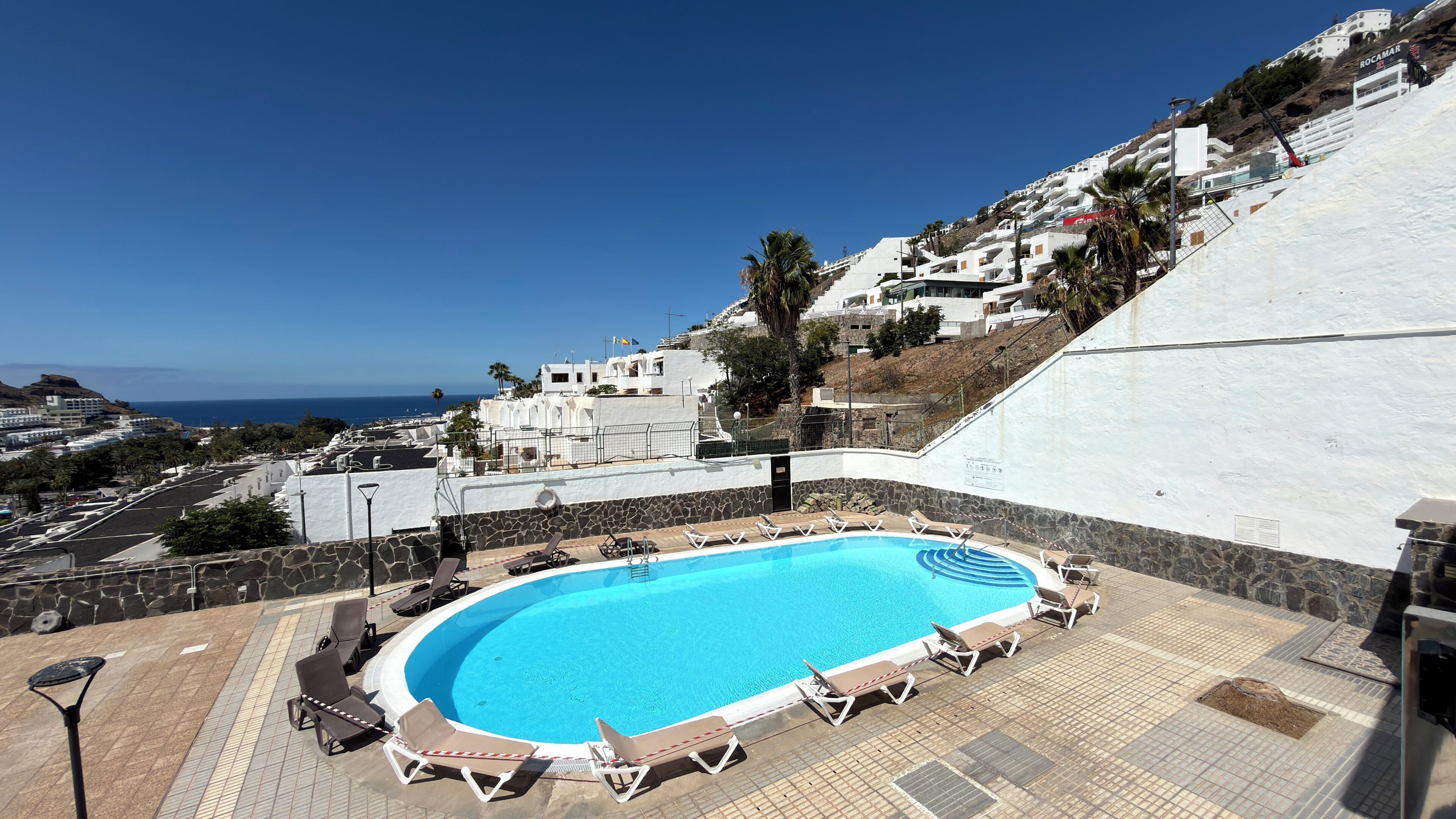 A swimming pool with lounge chairs, a stone wall, and a view of the ocean and white buildings. - Sunny Sea View Puerto Rico by Alquiler de Sueño (Mogan)