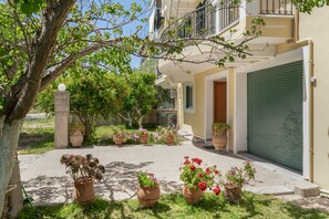 A building with a closed garage door, potted plants, and a tree in the foreground. - Jennifer's Apartments Lefkada – Comfortable Stay with City Views (Lefkada)