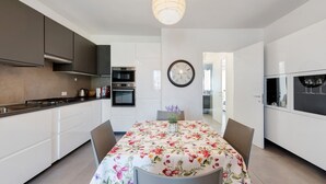 A modern kitchen with white cabinets, a dining table with a floral tablecloth, and a wall clock. - Apartment 'Caky's House' with Shared Terrace, Wi-Fi and Air Conditioning (Desenzano del Garda)