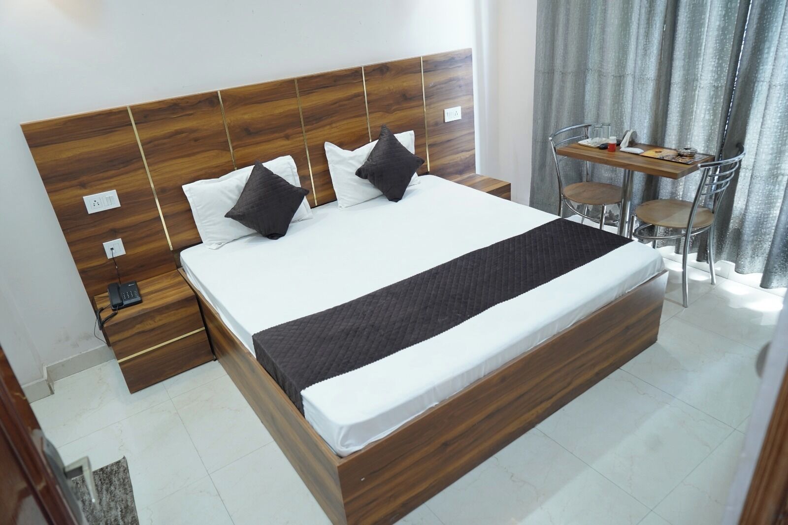 Una habitación de hotel con cabecera de madera, cama con ropa blanca, una manta de color negro y una pequeña mesa con sillas.