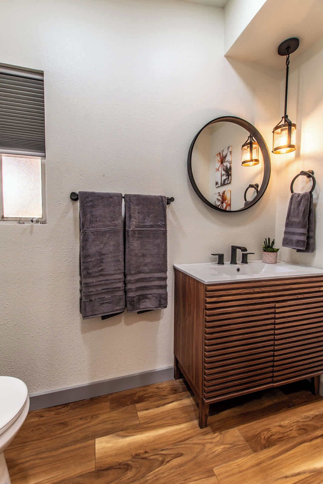 Un baño con tocador de madera, un espejo redondo y dos toallas colgadas en un perchero.