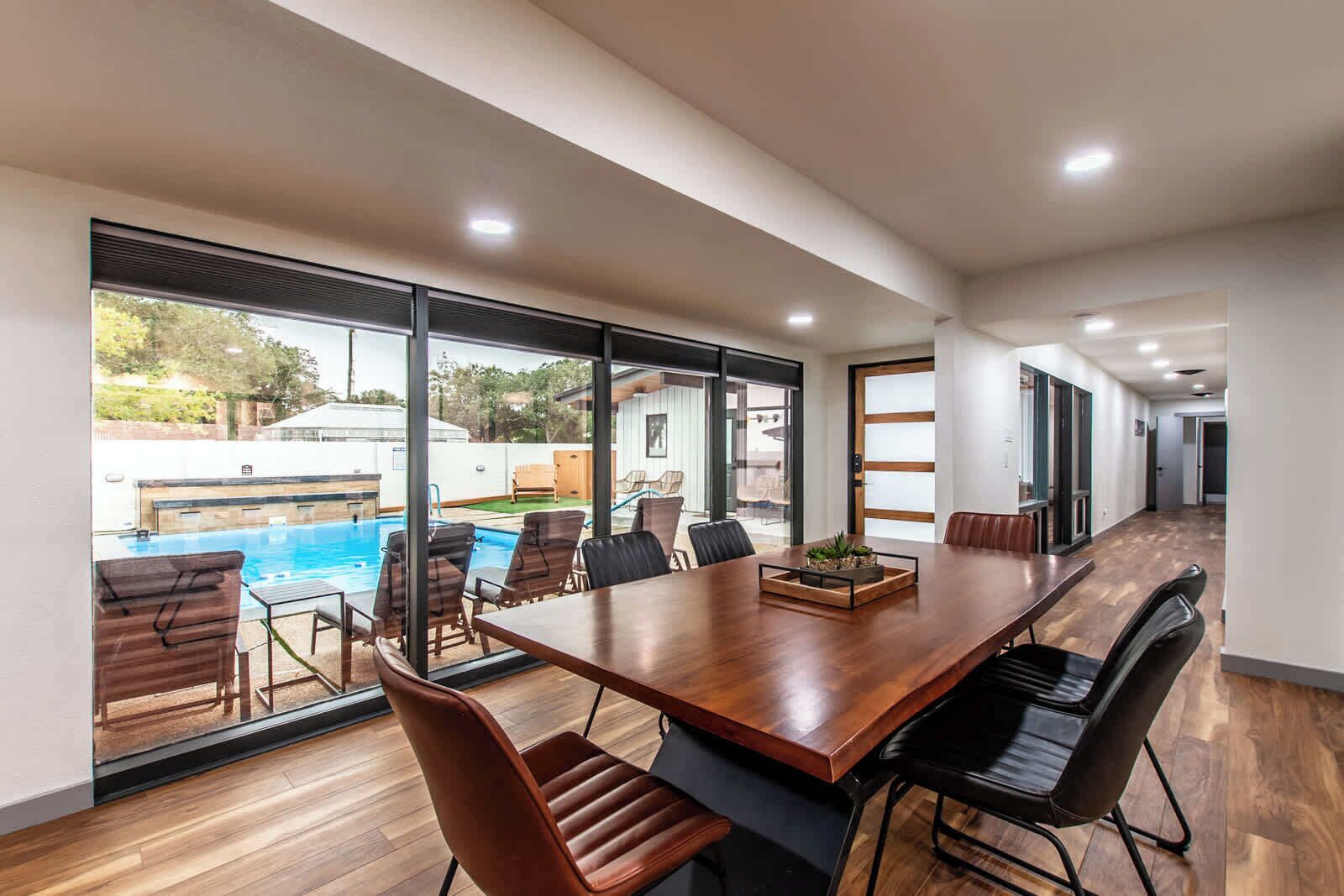 A spacious indoor area with a large wooden table and chairs, large windows offering a view of an outdoor swimming pool.