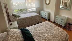 Una camera da letto con due letti, una ventola a soffitto, una finestra con persiane, un comò, uno specchio e un portalampada fissato al muro.