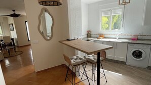 Una cucina con un tavolo in legno, due sgabelli da bar, un lampadario, una lavatrice e una finestra con persiane.