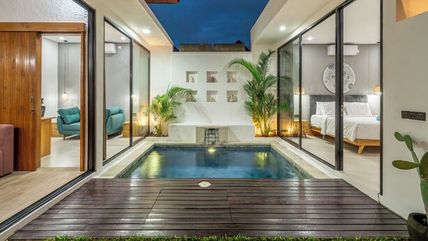 A modern indoor pool area with glass walls, a wooden deck, and a bed with white bedding. - Harmony Villas  (Denpasar)