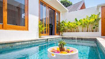 A swimming pool with a decorative island, a modern house with large windows, and a wall of tropical plants.