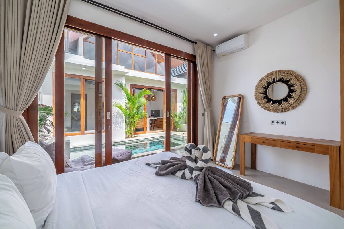 Un dormitorio con cama, mesita de noche, espejo y vista a una piscina.