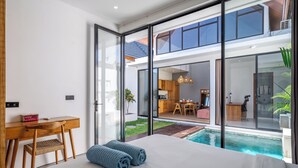 A modern bedroom with a large bed, a desk, and a view of a pool and outdoor area. - Harmony Villas  (Denpasar)