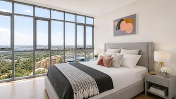 A bedroom with a large window, a bed with pillows, a bedside table with a lamp, and a painting on the wall.