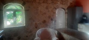 A room with a stone wall, a window with a view of greenery, a door, and a kitchenette area. - Traditional Stone House in Kerkira Argyrades (Argyrades Kerkira)