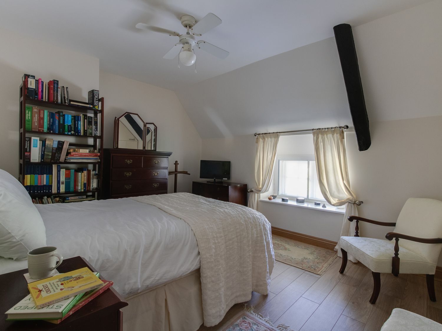 A bedroom with a bed, a bookshelf, a dresser, a TV, a window with curtains, a chair, and a ceiling fan.