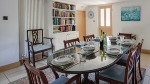 A dining area with a glass table, chairs, a bookshelf, and a cityscape painting. - Honeypot Cottage (Weymouth)