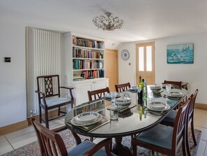 A dining area with a glass table, chairs, a bookshelf, and a cityscape painting. - Honeypot Cottage (Weymouth)