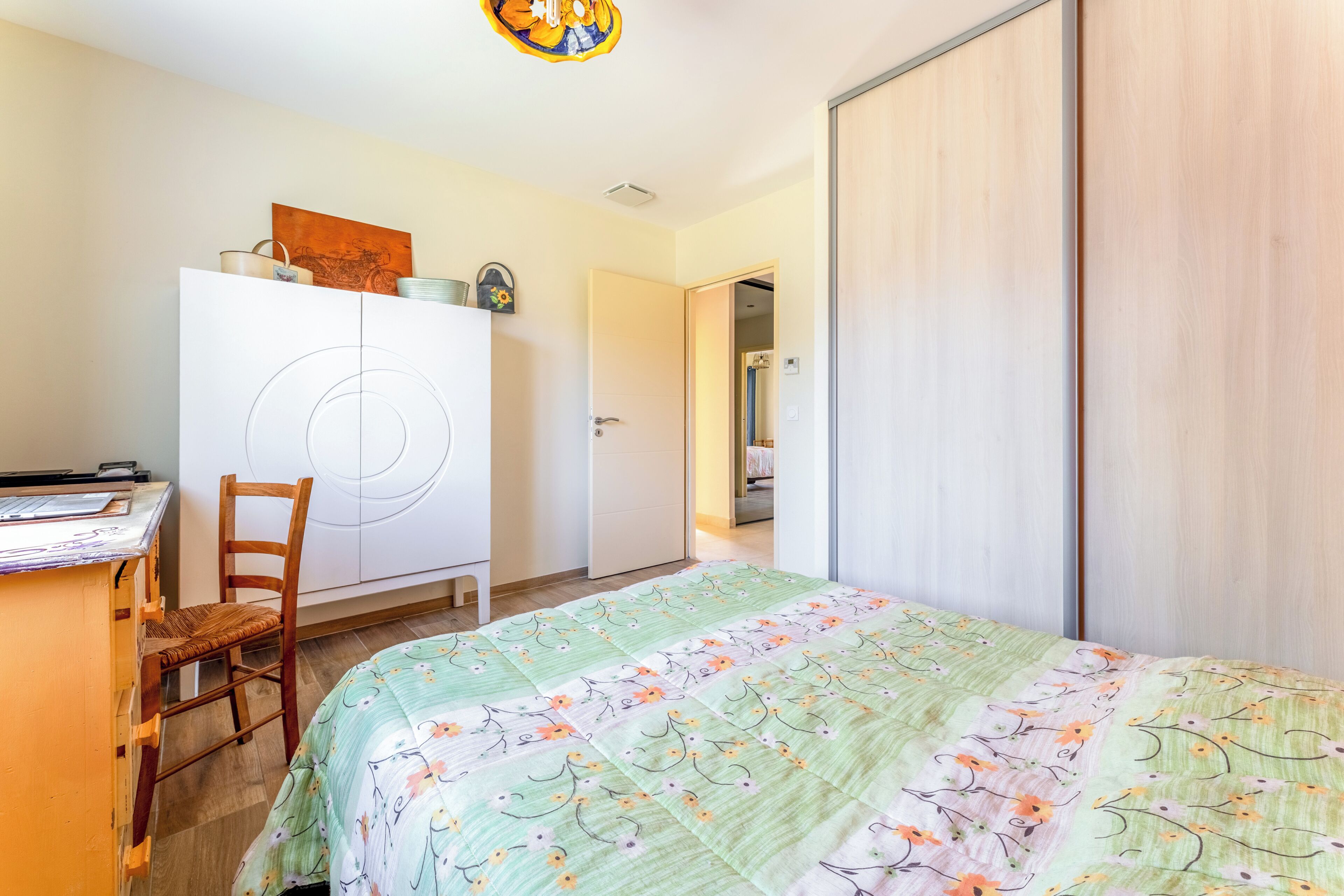 A bedroom with a bed, a desk, a chair, a wardrobe, and a door leading to another room.