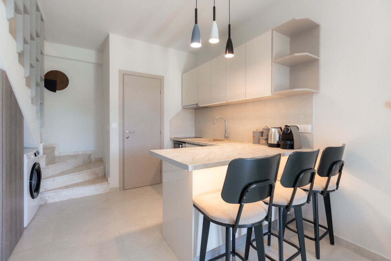 A modern kitchen with a central island, bar stools, and a staircase leading to another room.