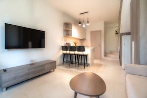 Un salon moderne dotĂ© dâun tĂ©lĂ©viseur Ă Ă©cran plat, dâun coin repas avec des chaises et une table, et dâune cuisine avec un plan de travail et des placards.