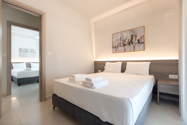Une chambre dâhĂŽtel avec un grand lit, une table de chevet, une commode, un tableau au mur et une porte donnant sur une autre piĂšce.