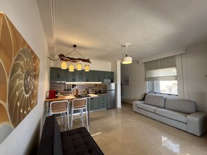 A modern living room with a sofa, a dining area with chairs, a kitchen with cabinets and appliances, and a large window with blinds. - Cali-Waves Apartment by RentalsPro - Halkidiki (Poligiros)