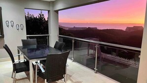 Un balcon doté d’une balustrade en verre, une table à manger avec des chaises, et une vue sur un coucher de soleil se déclinant sur un paysage.