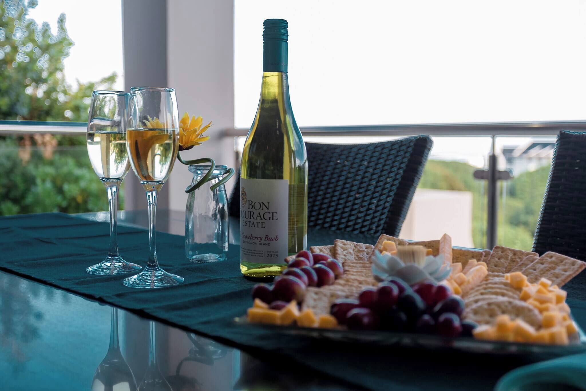 A table with two glasses of white wine, a bottle of wine, and a cheese platter with grapes and crackers.