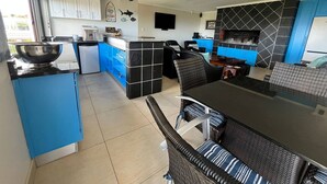 Une cuisine moderne avec des meubles bleus, un coin repas avec des meubles en osier et une cheminée recouverte de carreaux noirs.
