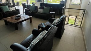 Un vaste espace de vie comprenant un grand canapé, une table basse et une table à manger. De nombreuses fenêtres offrent une vue sur la mer.