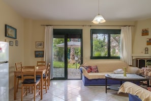 Ein Zimmer mit Esstisch, Stühlen, einer Couch, einem kleinen Tisch mit einer Vase und Blick ins Freie durch große Fenster.