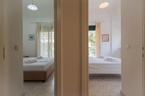 Ein Hotelzimmer mit zwei Betten, einem fensterbelaubten Fenster, einem Bild an der Wand und einer Tür zu einem weiteren Raum.