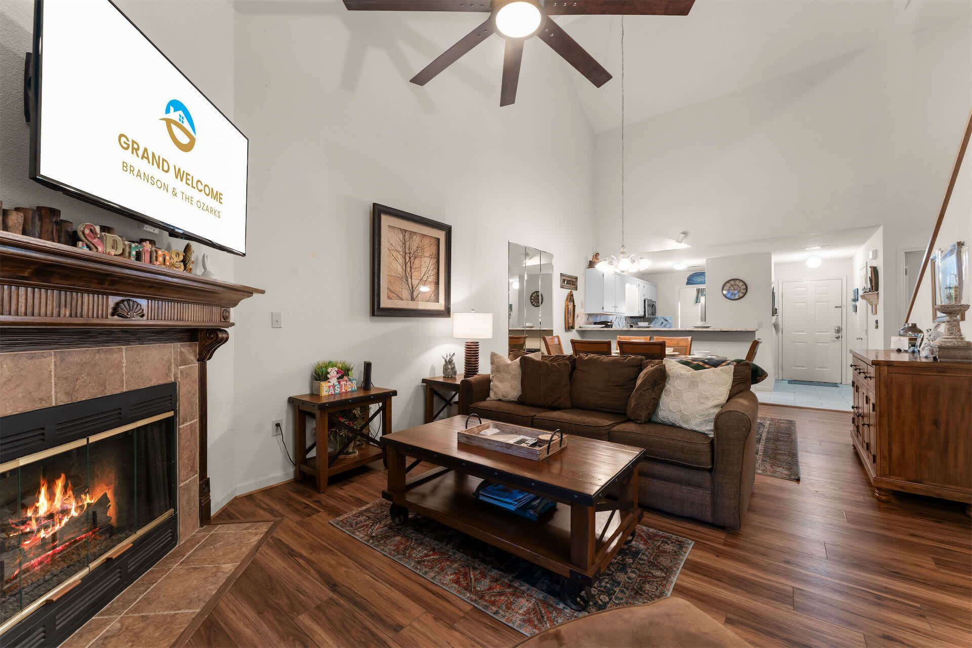 A living room with a fireplace, a ceiling fan, a sofa, a coffee table, and a kitchen in the background.