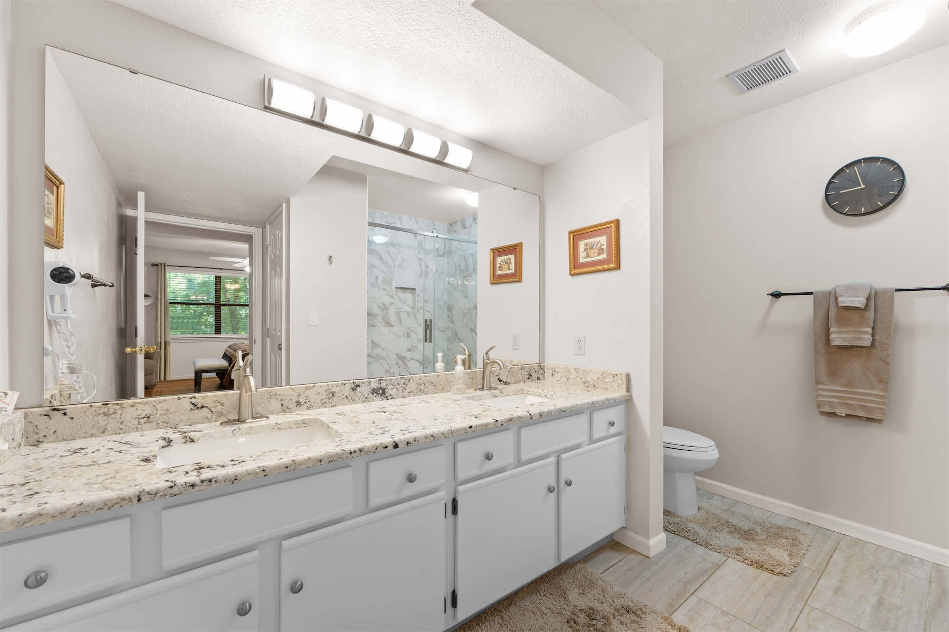 A bathroom with a large mirror, a sink with a marble countertop, a toilet, and a clock on the wall.