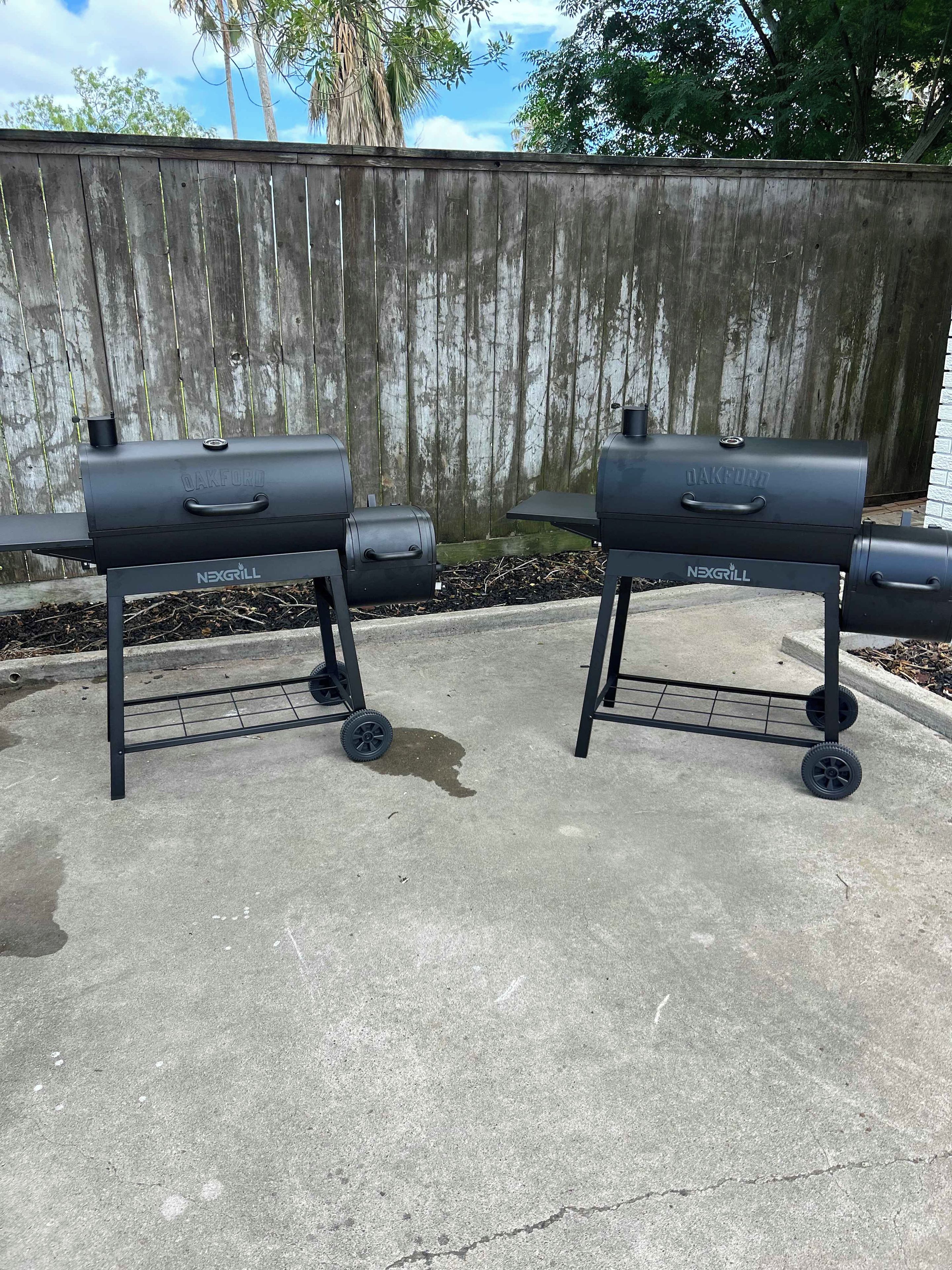 Dos parrillas para exterior negras con ruedas, con las marcas 'NEXGRILL' y 'OAKFORD', sobre un patio de concreto con una cerca de madera al fondo.