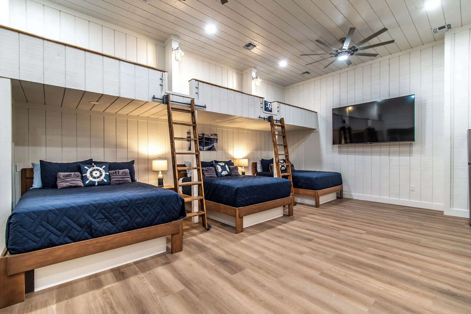 Une pièce avec trois lits superposés, un plancher en bois, des murs recouverts de panneaux blancs, un ventilateur de plafond et un téléviseur à écran plat fixé au mur.