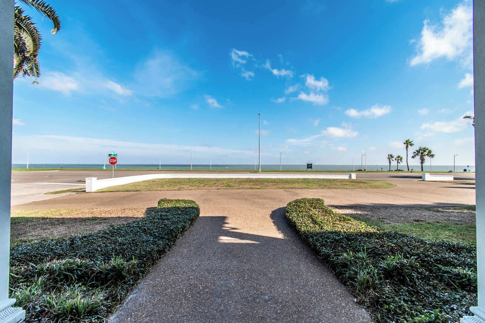 Un camino pavimentado que lleva a una señal de parada en una ubicación junto a la playa, con palmeras y cielo despejado.