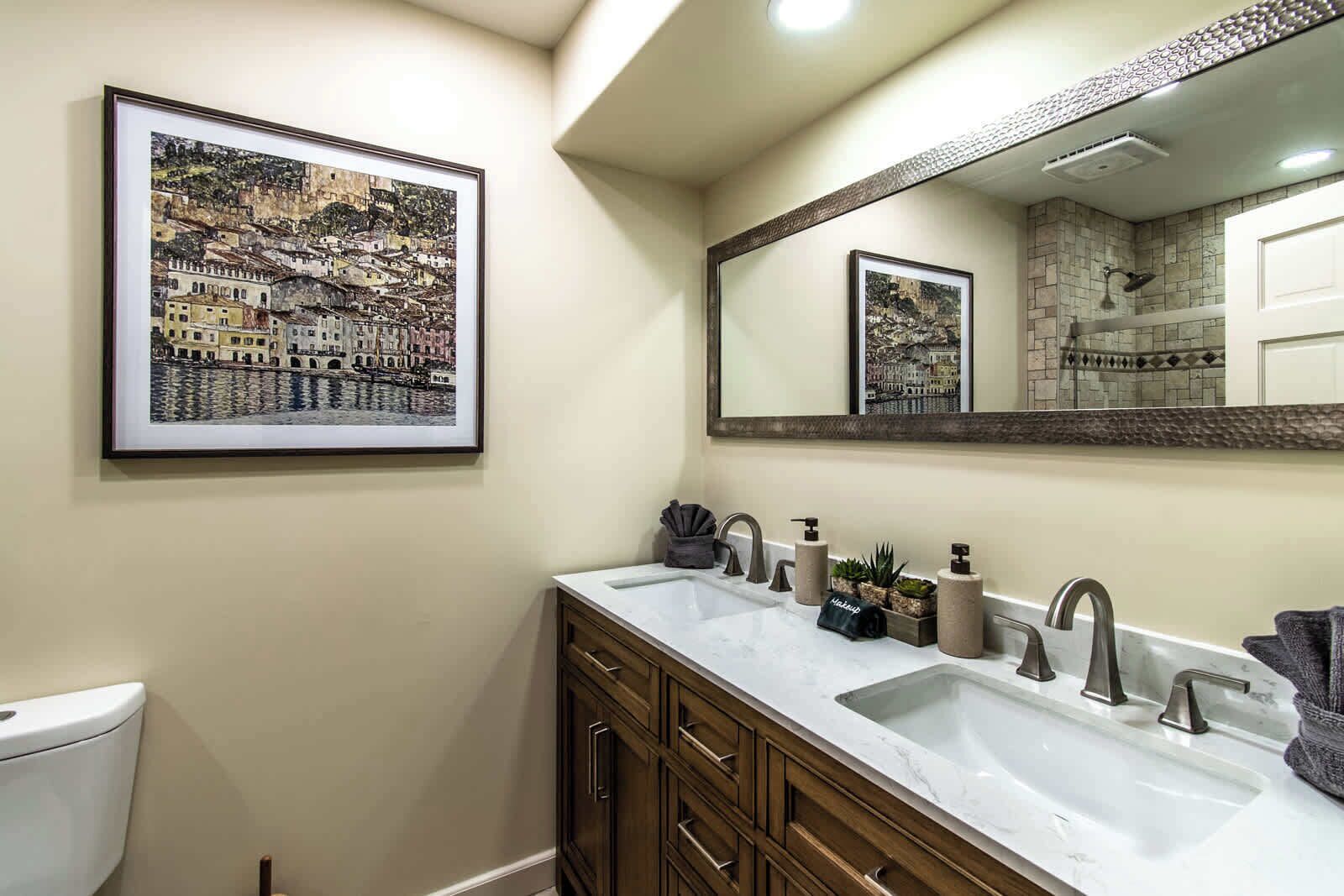 A bathroom with a large mirror, a sink with two faucets, and a toilet. There are framed pictures on the wall.