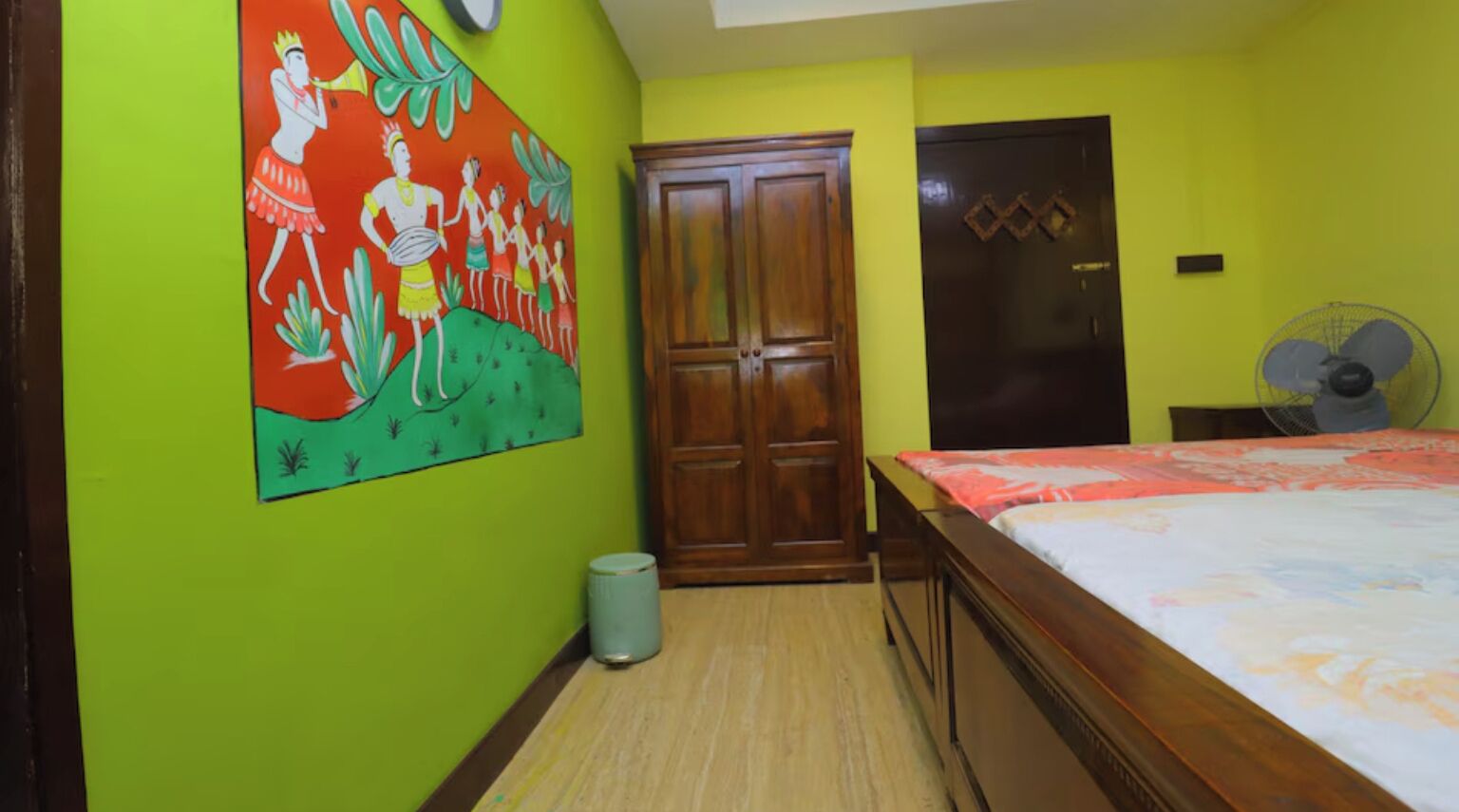 A room with a bed, a wooden wardrobe, a wall-mounted fan, and a trash bin.