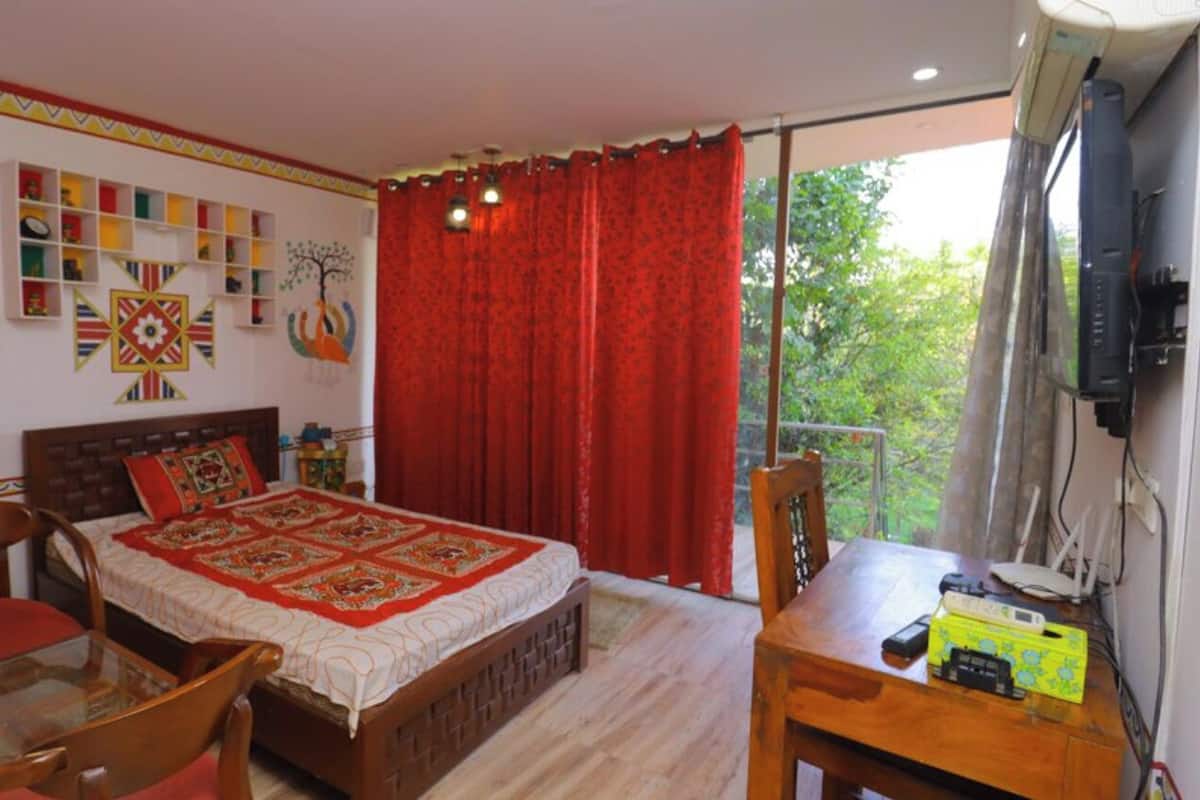 A bedroom with a bed, a table, a chair, a TV, and a balcony with red curtains.