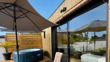A modern outdoor seating area with a table set for two, a blue hot tub, and a glass-enclosed room with a bed.