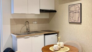 A compact kitchen with white cabinets, a built-in dishwasher, and a small refrigerator. There is a round wooden table with two chairs, a wall-mounted air conditioner, and a framed abstract art piece. - Cozy lodge with a private hot tub and air conditioning (La Trinité-Surzur)