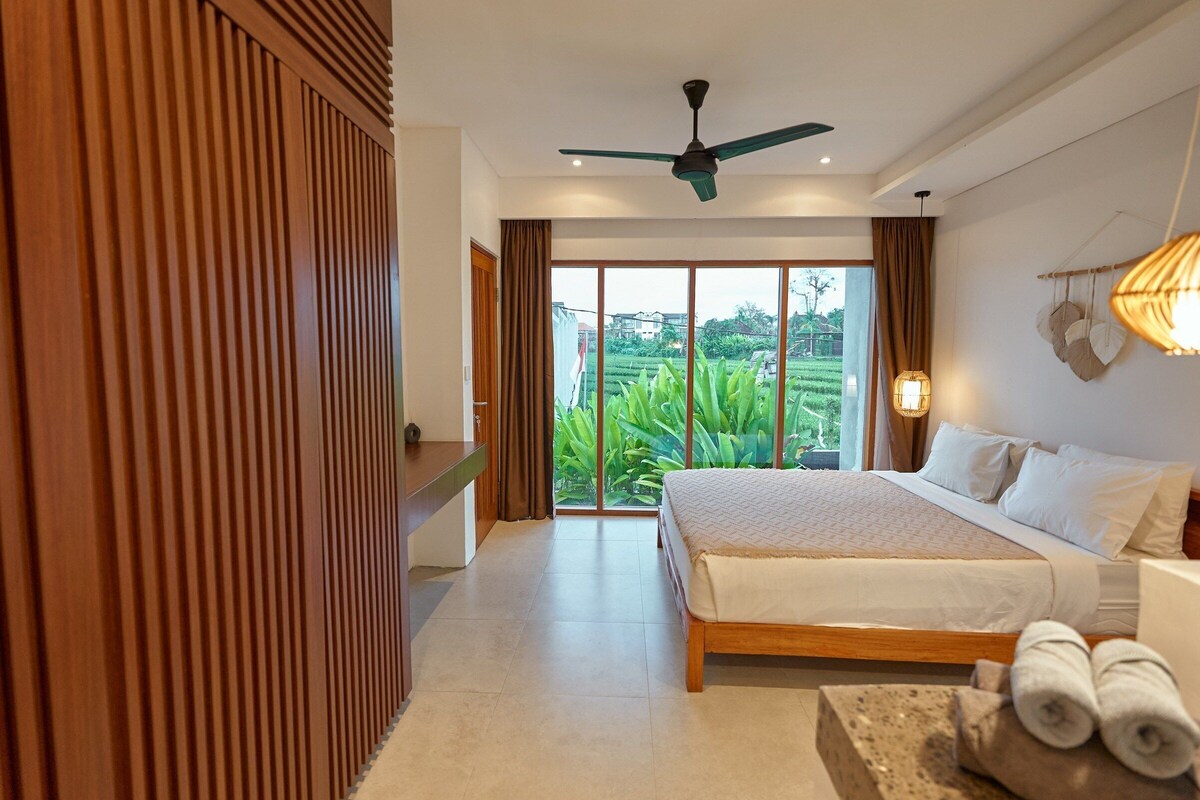 Un dormitorio con cama, ventilador de techo, vista a zonas verdes y pared de paneles de madera.