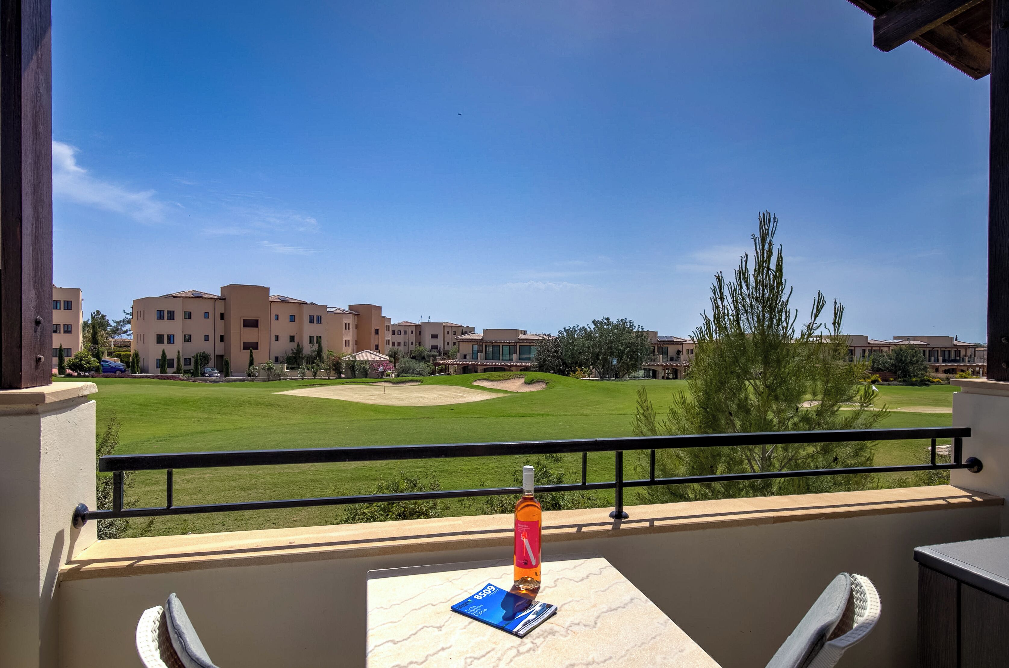 A balcony with a table, a bottle, and a card, overlooking a golf course and buildings.