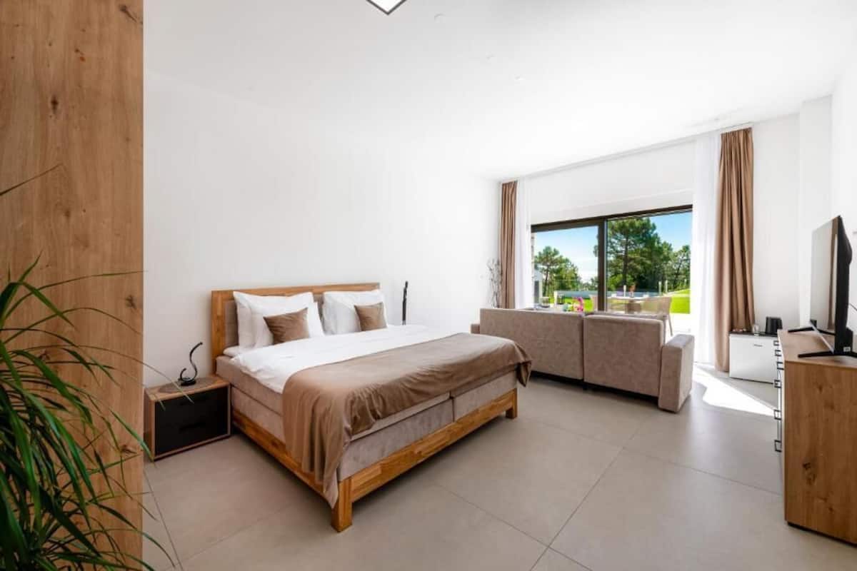 Un dormitorio moderno con una cama grande, un sofá, un televisor y un ventanal con vistas a zonas verdes.